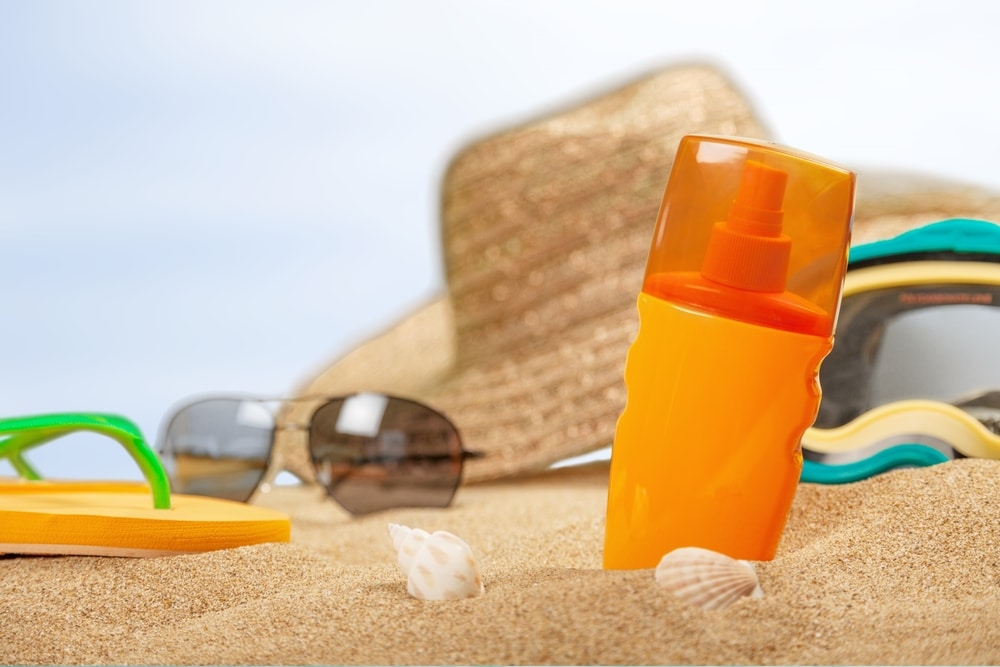 Sunscreen in sand, flip flop, sunglasses, and hat in background