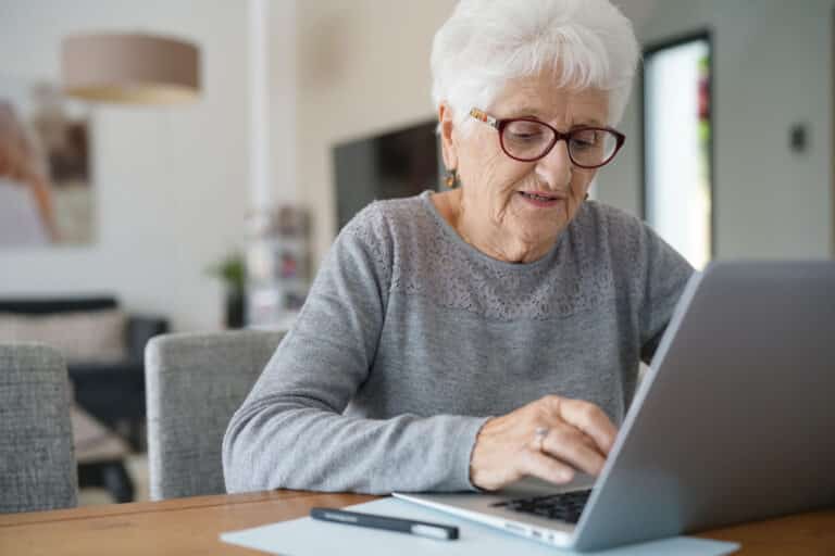 Senior woman typing on laptop, notebook beside her