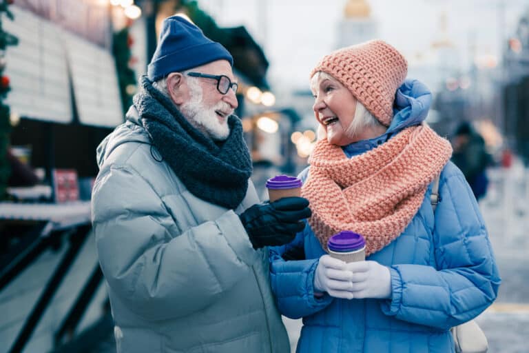 Two smiling seniors dressed warmly and holding hot drinks outside in winter