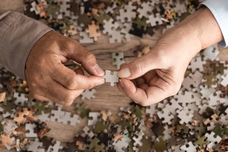 Two hands holding piece of jigsaw puzzle above table with lots of puzzle pieces