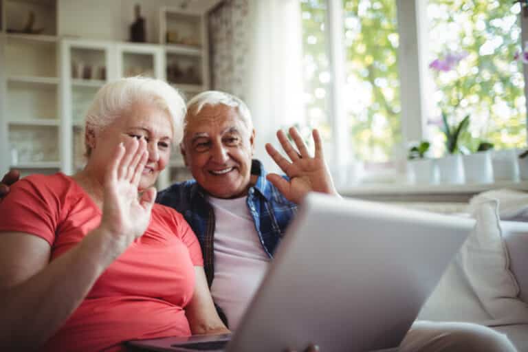 Two seniors smiling and waving at their laptop, video chatting